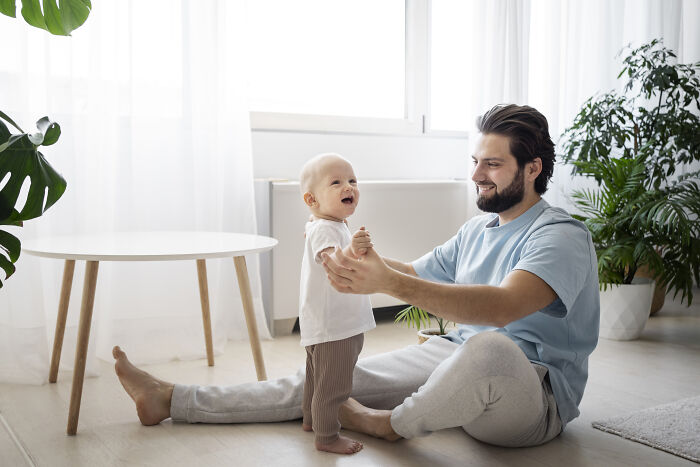 Father smiling and playing with baby standing between legs in bright room with plants, illustrating baby names concept.