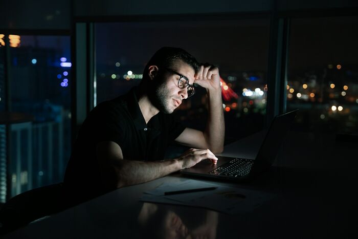 Young man wearing glasses working late on laptop, showing behaviors that may indicate someone is insecure.