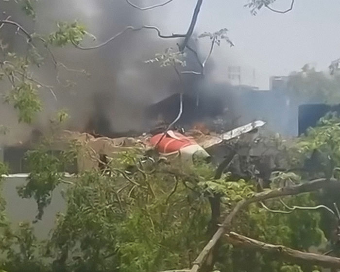 Wreckage of Air India plane crash with smoke rising, surrounded by trees and nearby buildings visible in background.