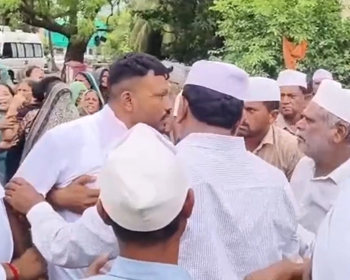 Sole survivor of Air India crash embraced by men in white caps during emotional gathering outdoors near trees and vehicles.