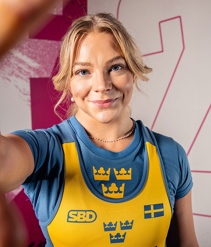 Female powerlifter wearing Swedish singlet smiling in gym, related to viral video of woman urinating during powerlifting competition. Female powerlifter wearing Swedish singlet smiling in gym, related to viral video of woman urinating during powerlifting competition.