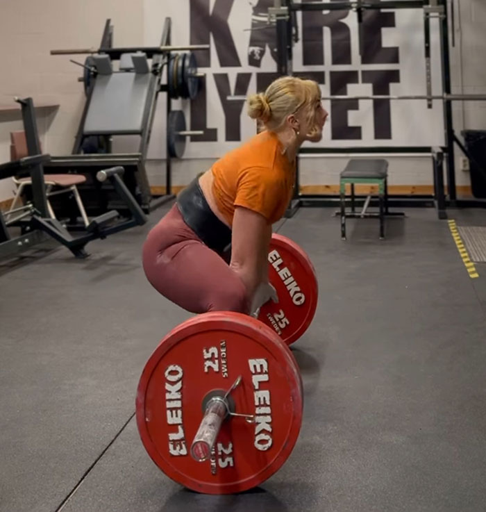 Woman powerlifting in gym wearing orange shirt and pink leggings, preparing to lift Eleiko barbell weights. Woman powerlifting in gym wearing orange shirt and pink leggings, preparing to lift Eleiko barbell weights.