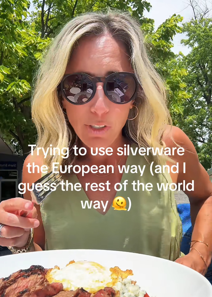 Woman wearing sunglasses outdoors holding a fork, attempting European style eating with cutlery against a meal backdrop.