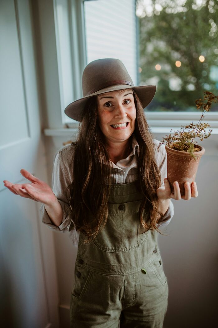 Woman wearing a hat and overalls holding a potted plant with a hesitant expression showing insecure behaviors.