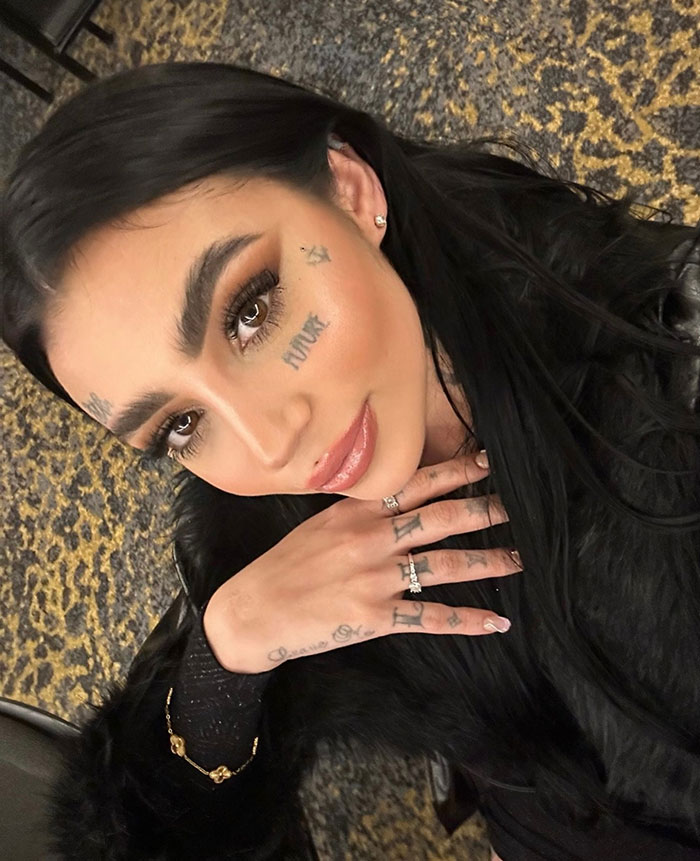 Young woman with face and hand tattoos wearing a black fur jacket, highlighting plastic surgery and tragedy keywords.