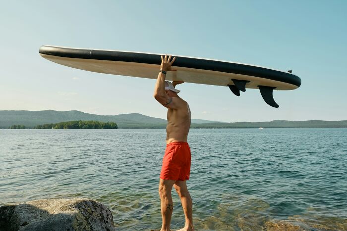 Man in red shorts holding a paddleboard overhead by the lake, enjoying a priceless reward from a simple dollar.