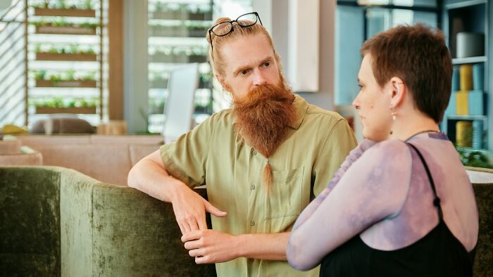 Man with long red beard listens attentively to woman in casual conversation about pregnancy and twins indoors.