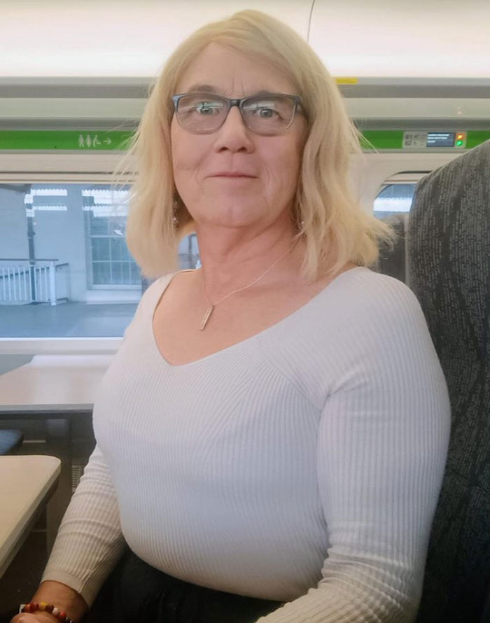Trans woman with blonde hair and glasses sitting on a train, related to trans woman swims topless competition protest.