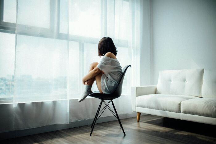 Person sitting alone on a chair by a window in a minimalist room, reflecting on the current situation and opinions.