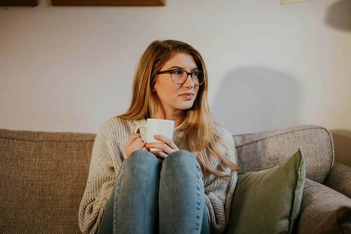 Young mom sitting on couch holding a white mug, looking thoughtful in a cozy home setting. Young mom sitting on couch holding a white mug, looking thoughtful in a cozy home setting.
