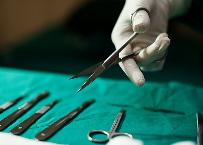 Autopsy doctor wearing gloves holding scissors over surgical tools on a green cloth during autopsy procedure.