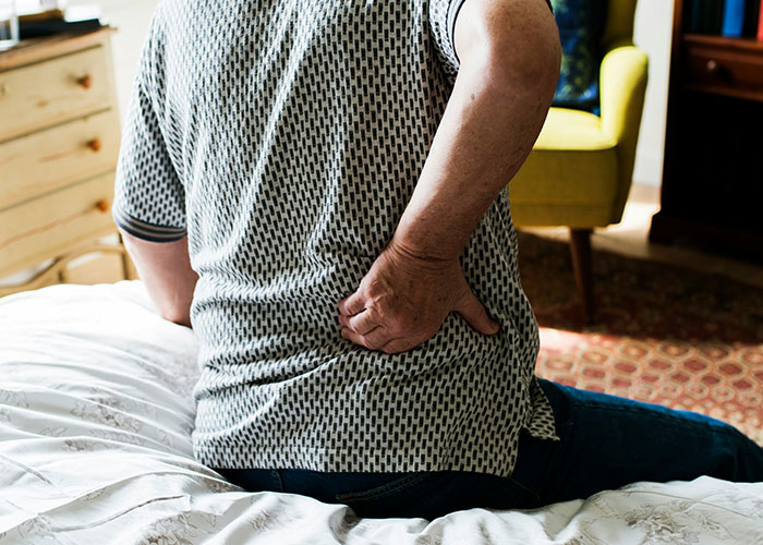 Person sitting on bed holding lower back in pain, illustrating strange findings autopsy doctors have discovered on the job.