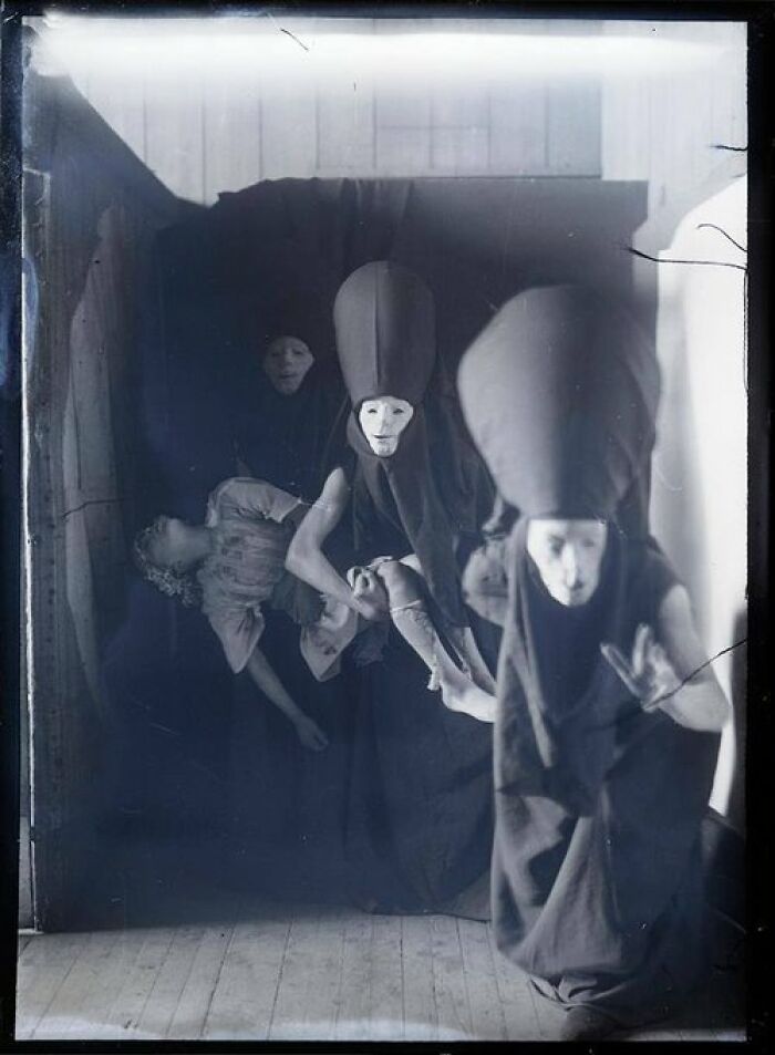 Creepy and unsettling vintage photo of three figures in dark robes and masks holding a limp doll in a dim room.