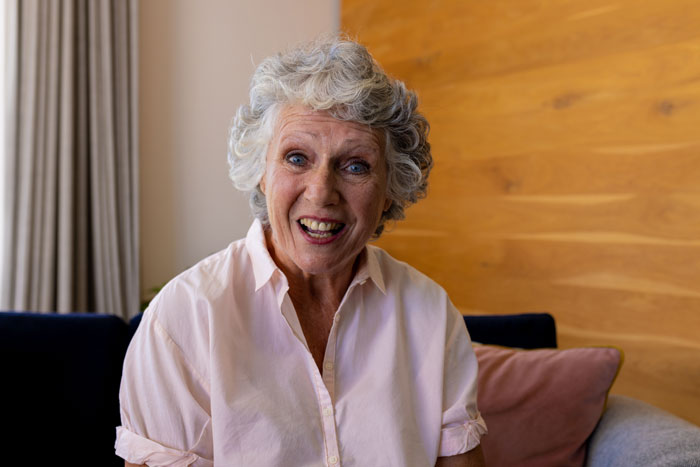 Elderly woman with curly gray hair sitting indoors, portraying a concerned mother-in-law in a family conflict scenario.