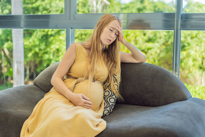 Pregnant woman in a yellow dress looking stressed while sitting on a gray sofa at a gender reveal party.