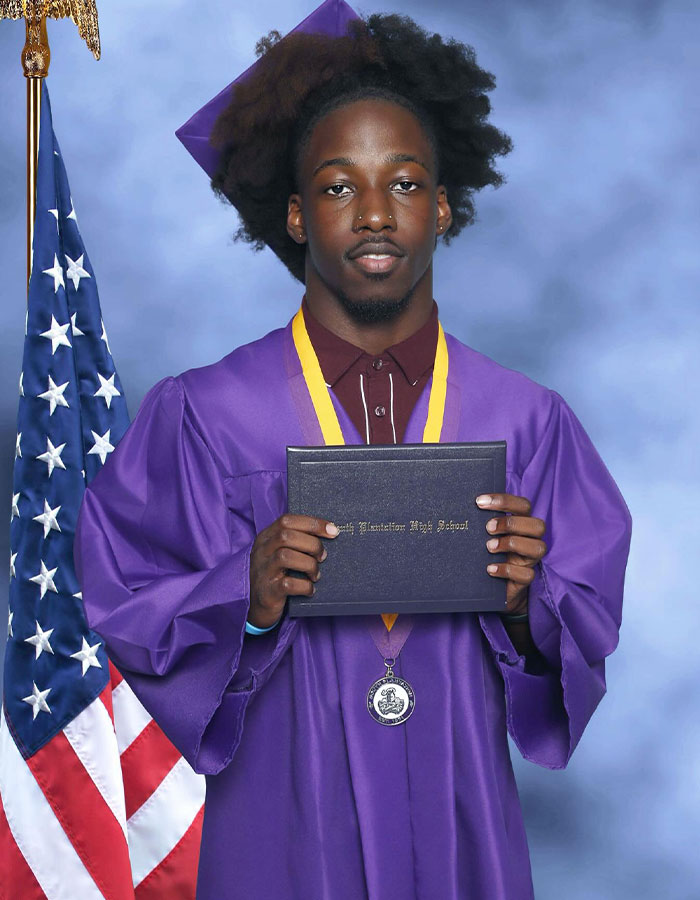 Florida high school grad wearing purple cap and gown, holding diploma, standing in front of American flag during graduation.