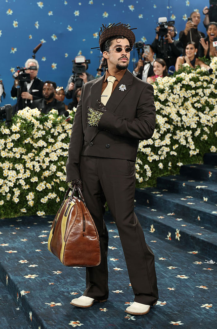 Bad Bunny posing in a brown suit and hat with a large leather bag at Paris Fashion Week event.