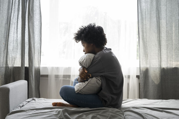 Person sitting on a bed near window, hugging pillow, reflecting on bad things in life in a quiet, dimly lit room.