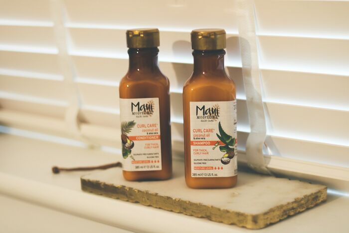 Two Maui Moisture hair care bottles with curl care shampoo and conditioner on a windowsill with white blinds.