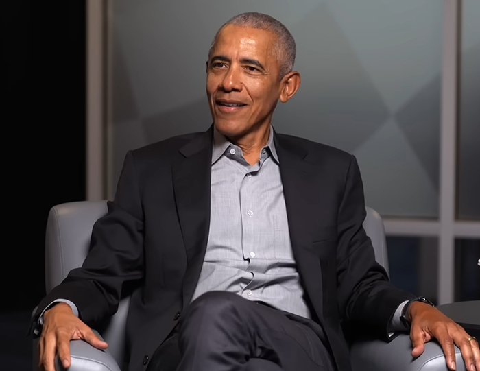 Barack Obama sitting in a chair, dressed in a black blazer and gray shirt, during a formal interview setting. Barack Obama sitting in a chair, dressed in a black blazer and gray shirt, during a formal interview setting.