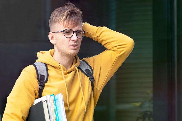 High school student in yellow hoodie looks confused while holding books, illustrating lack of knowledge educators warn about. High school student in yellow hoodie looks confused while holding books, illustrating lack of knowledge educators warn about.