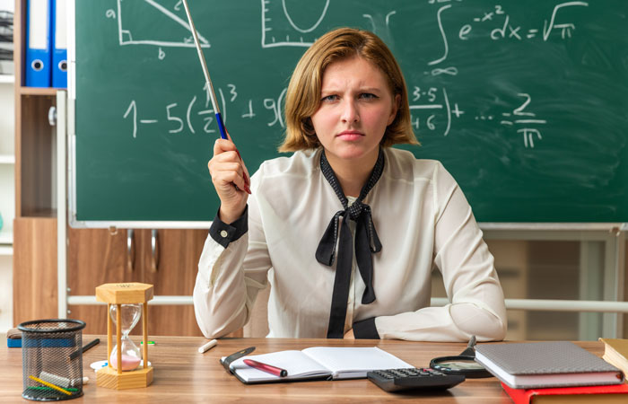 Teacher looking frustrated in classroom with chalkboard behind her, highlighting educators’ concerns over high schoolers' knowledge gaps. Teacher looking frustrated in classroom with chalkboard behind her, highlighting educators’ concerns over high schoolers' knowledge gaps.
