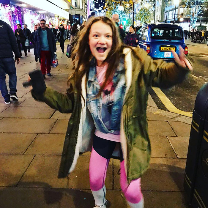Young woman wearing a green coat and pink leggings on a busy city sidewalk, relating to Greta Thunberg's singer sister story.
