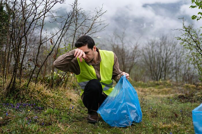 Man wearing a safety vest holding a blue trash bag, picking up litter outdoors in a natural setting with trees and fog.