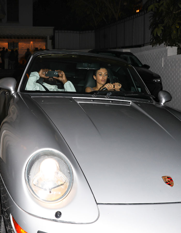Bianca Censori driving a grey Porsche at night with a passenger using a smartphone inside the car.