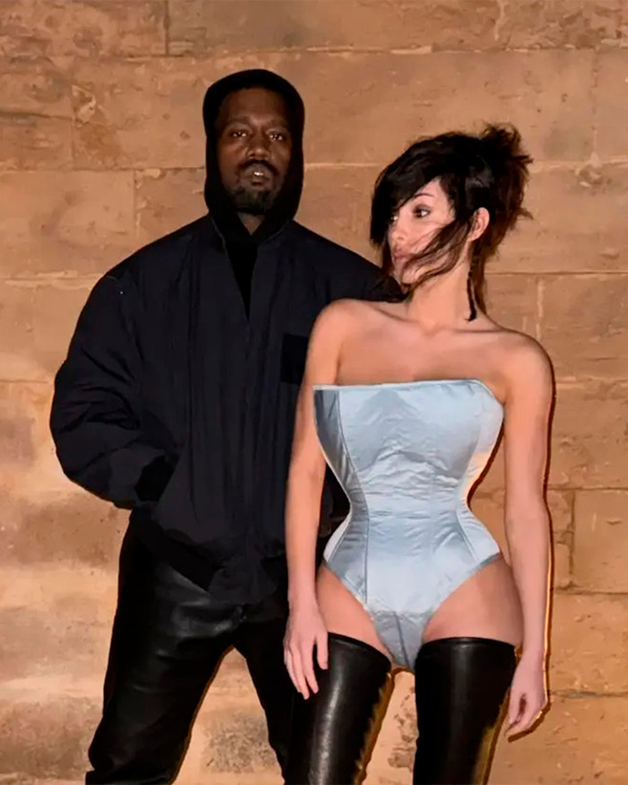 Kanye West standing next to a woman in a corset and thigh-high boots against a stone wall at night.