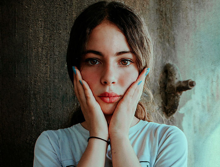 Young woman with hands on cheeks, illustrating human body flaws and vulnerabilities in natural light setting.