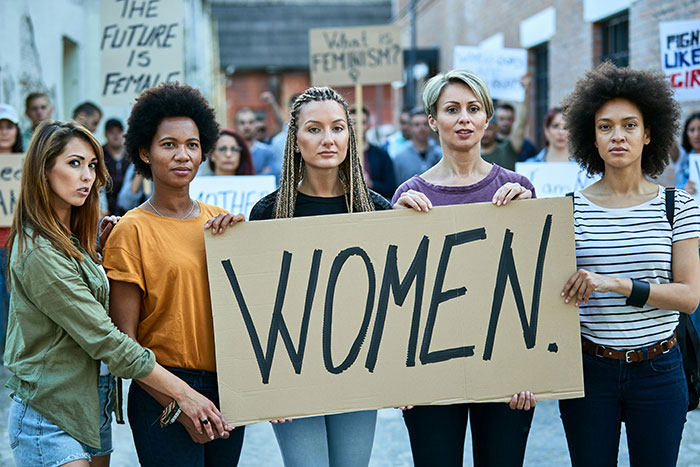 Group of diverse women holding a sign saying WOMEN during a protest, representing voices in the biggest scandal in human history.