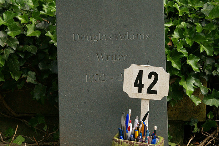 Gravestone of Douglas Adams surrounded by pens and a sign with number 42, symbolizing cultural references and historical scandals.