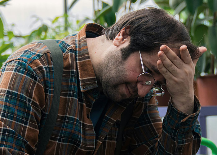 Man frustrated over kitchen renovation problems, wearing glasses and plaid shirt, sitting indoors with plants in background