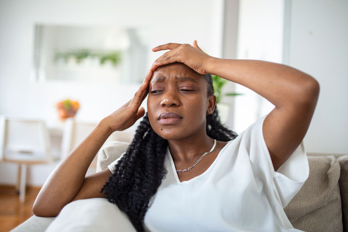 Teacher sitting on a couch with a pained expression, holding her head, stressed from paying family&rsquo;s bills for years.