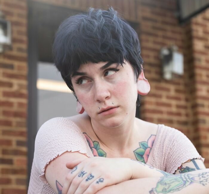 Young person with tattoos and ear gauges sitting outdoors, expressing thoughtful and unsettled emotions on current situation.