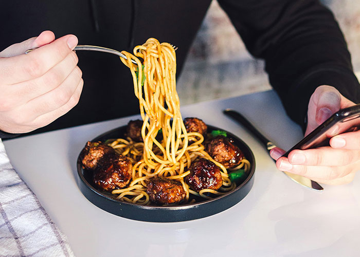 Person eating lunch with noodles and meatballs while looking at a phone during a meeting sacrifice break Person eating lunch with noodles and meatballs while looking at a phone during a meeting sacrifice break