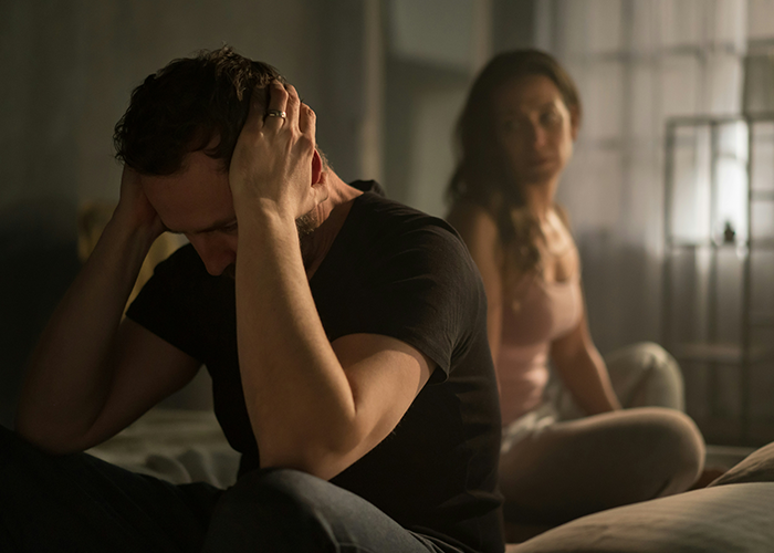 Man stressed and holding his head on bed while girlfriend looks on, illustrating micro-cheating conflict in a relationship.