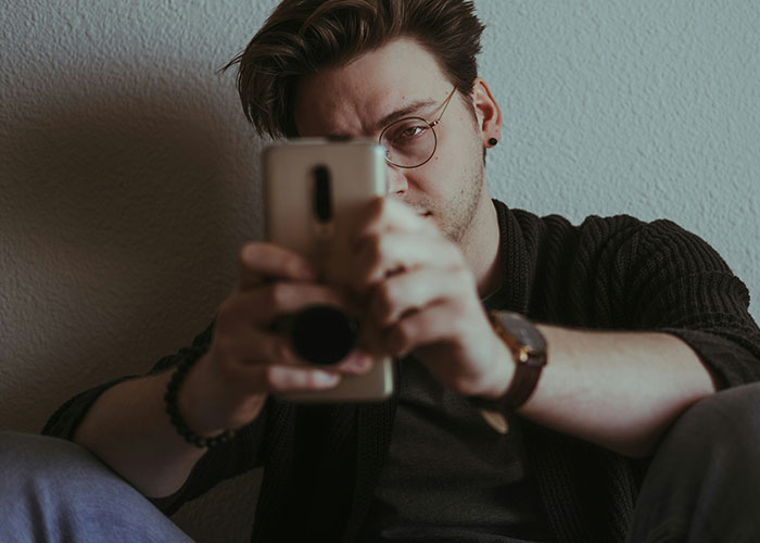 Man wearing glasses takes photo on smartphone, relating to guy finding old photo on girlfriend&rsquo;s chest and micro-cheating.