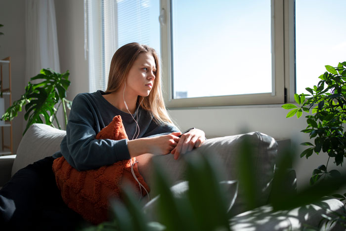 Young woman sitting on couch looking shocked and contemplative, reflecting on being called a downgrade by boyfriend&rsquo;s mom.