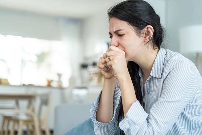 Young woman coughing and covering her mouth, showing discomfort while sitting in a bright home setting.