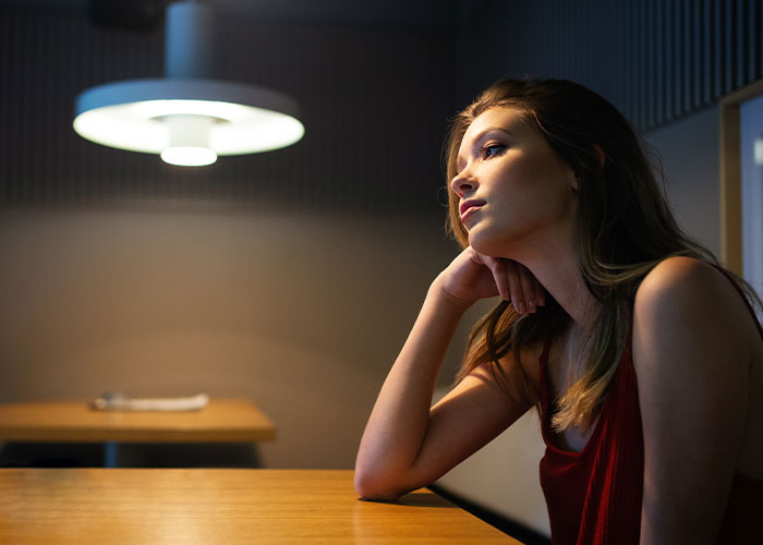 Young woman in a red top sitting alone at a table, refusing to leave party after boyfriend and friends demand privacy