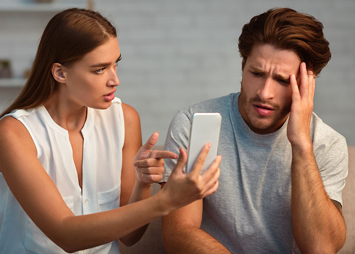 Young woman upset while showing phone to her friend who looks frustrated about his girlfriend not allowing them to sleep in the same bed