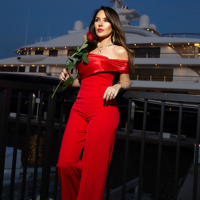 Vanessa Villela in a red jumpsuit holding a rose, standing by a railing with a yacht in the background at dusk.