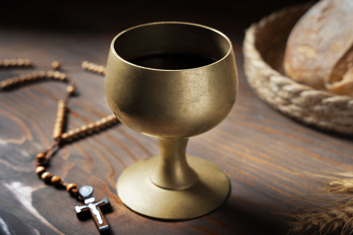 A brass chalice filled with red wine next to a wooden rosary on a rustic table depicting fictional things.