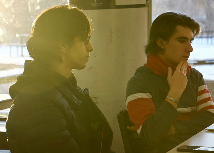 Two young men in casual clothes sitting indoors, reflecting the theme of breaking guy or girl code decisions.