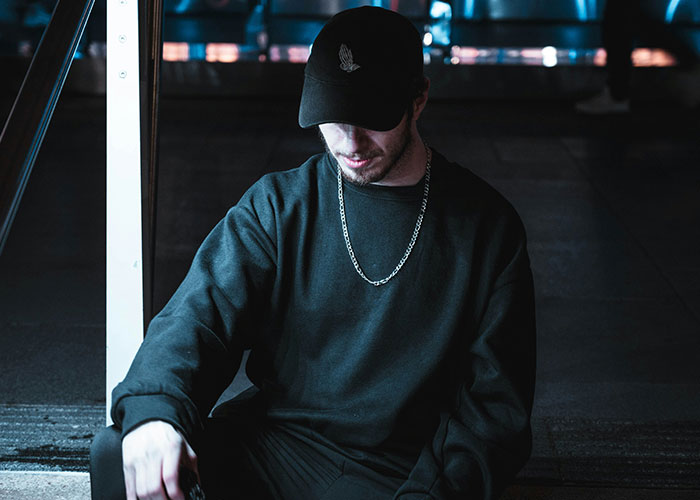 Young man wearing black cap and chain necklace sitting alone, capturing a moment breaking guy or girl code.