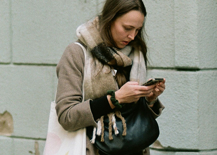 Woman breaking guy or girl code, focused on her phone while walking outdoors with scarf and coat in urban setting.