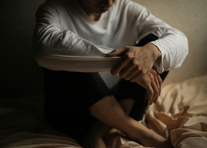 Person sitting on a bed in dim light, wearing a white long-sleeve shirt, reflecting on breaking guy or girl code choices.