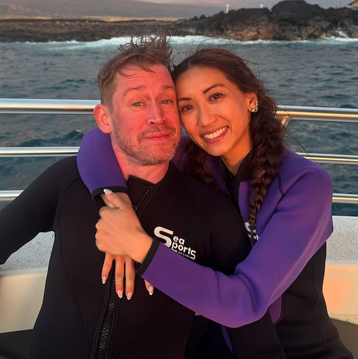 Couple wearing wetsuits hugging on a boat with ocean and rocky shoreline in the background at sunset.